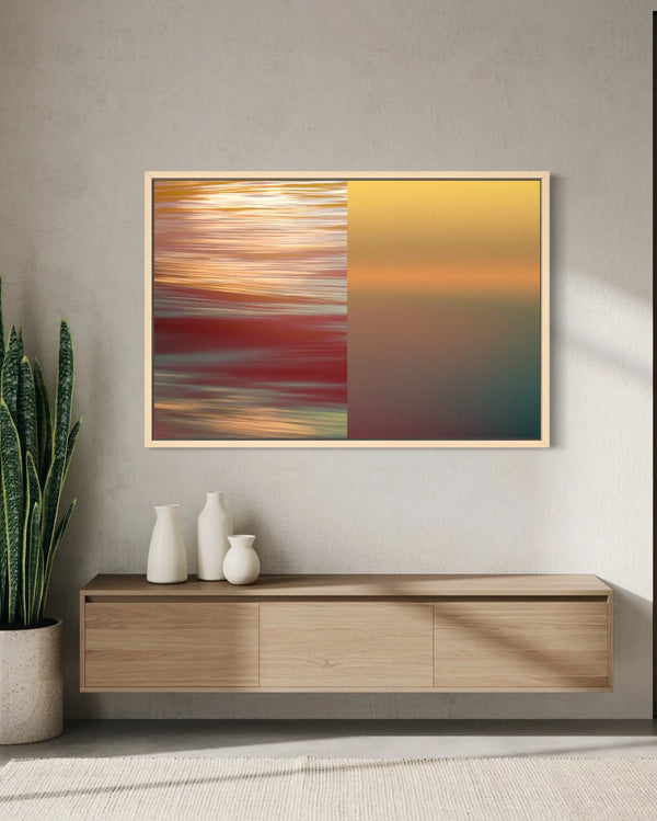 A framed abstract wall art print hangs above a light wood console table in a modern interior. The artwork is split into two vertical sections: the left side shows blurred streaks of red, orange, and gold, while the right side shows a smooth gradient from yellow to teal.