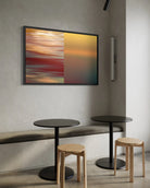 A framed abstract art print featuring a split design of orange, red, and yellow motion-blur on the left, and a smooth yellow-to-teal gradient on the right, displayed on a textured gray wall above a bench and tables.