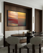 A framed abstract wall art print with a split red-orange and yellow design hangs on a dark wood-paneled wall above a dining area with upholstered chairs and a dark table.