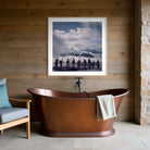 A framed square art print depicting a dramatic cloudy sky and silhouetted figures over water, displayed on a wood-paneled wall above a copper freestanding bathtub in a bathroom setting.