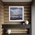 A framed square art print depicting a scenic mountain landscape with silhouetted figures, displayed on a rustic wood-paneled wall above a dark console table with decorative vases.
