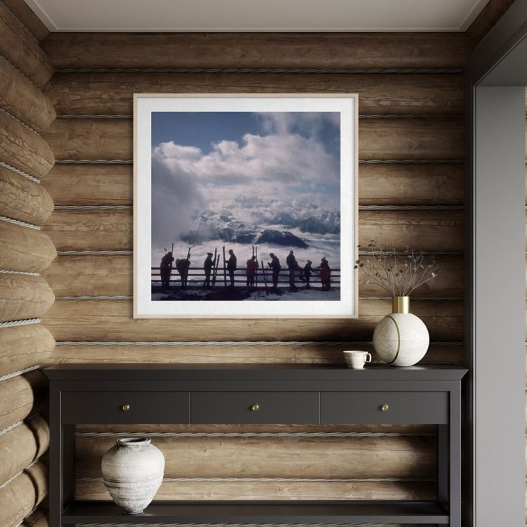 A framed square art print depicting a scenic mountain landscape with silhouetted figures, displayed on a rustic wood-paneled wall above a dark console table with decorative vases.