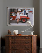 A framed art print depicting a vintage scene with a red race car and people, displayed on a dark gray wall above a brown woven dresser with a silver desk lamp.