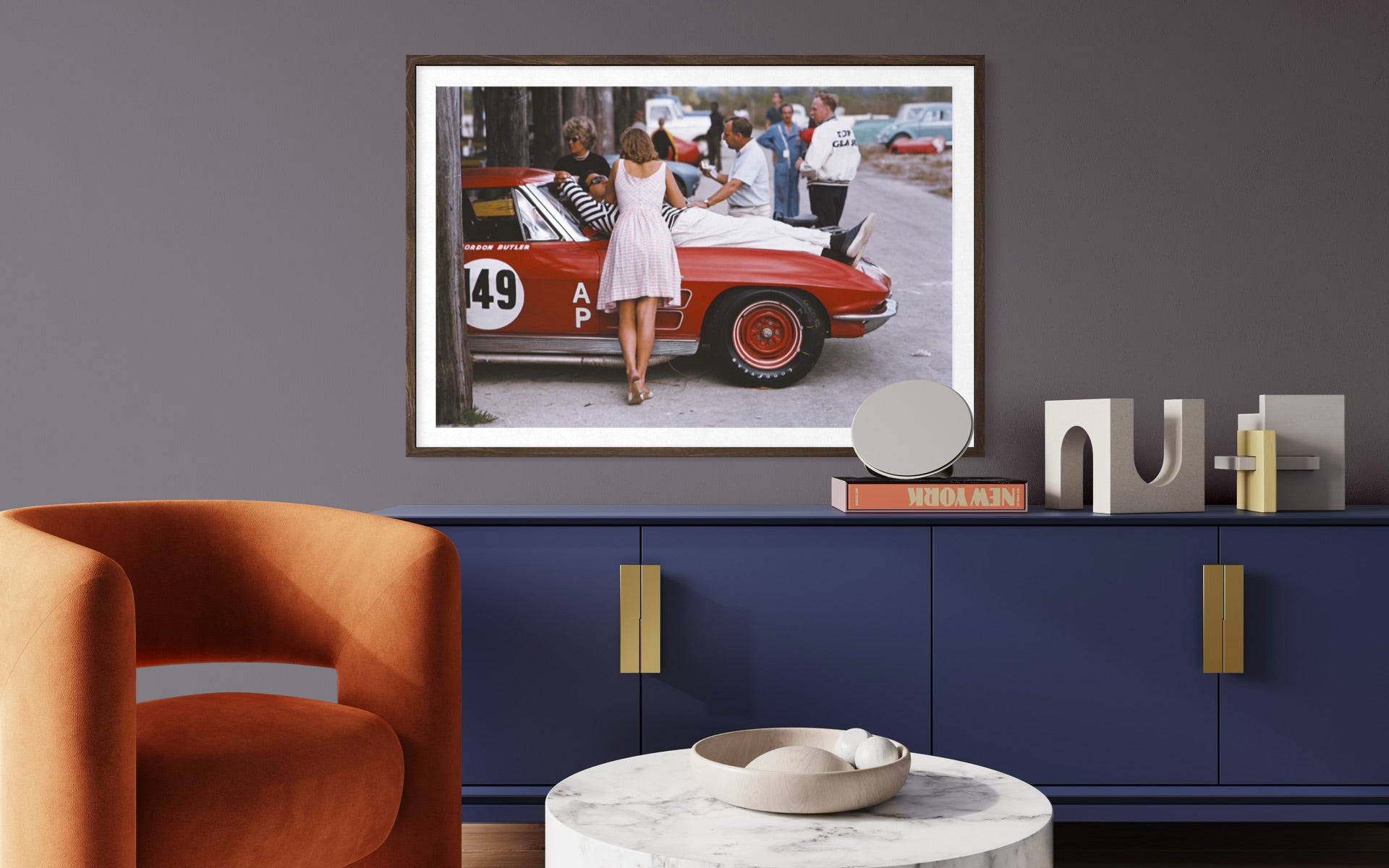 A framed art print depicting a vintage red sports car scene with a woman in a white dress, displayed on a dark gray wall above a blue cabinet in a modern living room setting.