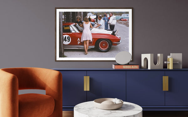 A framed art print depicting a vintage red sports car scene with a woman in a white dress, displayed on a dark gray wall above a blue cabinet in a modern living room setting.