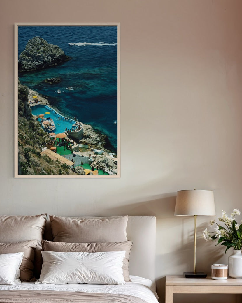 A vertical framed art print depicting a scenic coastal cove with turquoise water, rocky cliffs, and a beach, displayed on a beige wall above a bed in a modern bedroom setting.