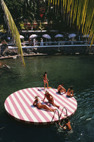 A large circular floating platform in a pool, featuring bold pink and white diagonal stripes. Several people are relaxing and standing on the platform, with palm leaves hanging over the scene and a resort-style background with umbrellas and lounge chairs.