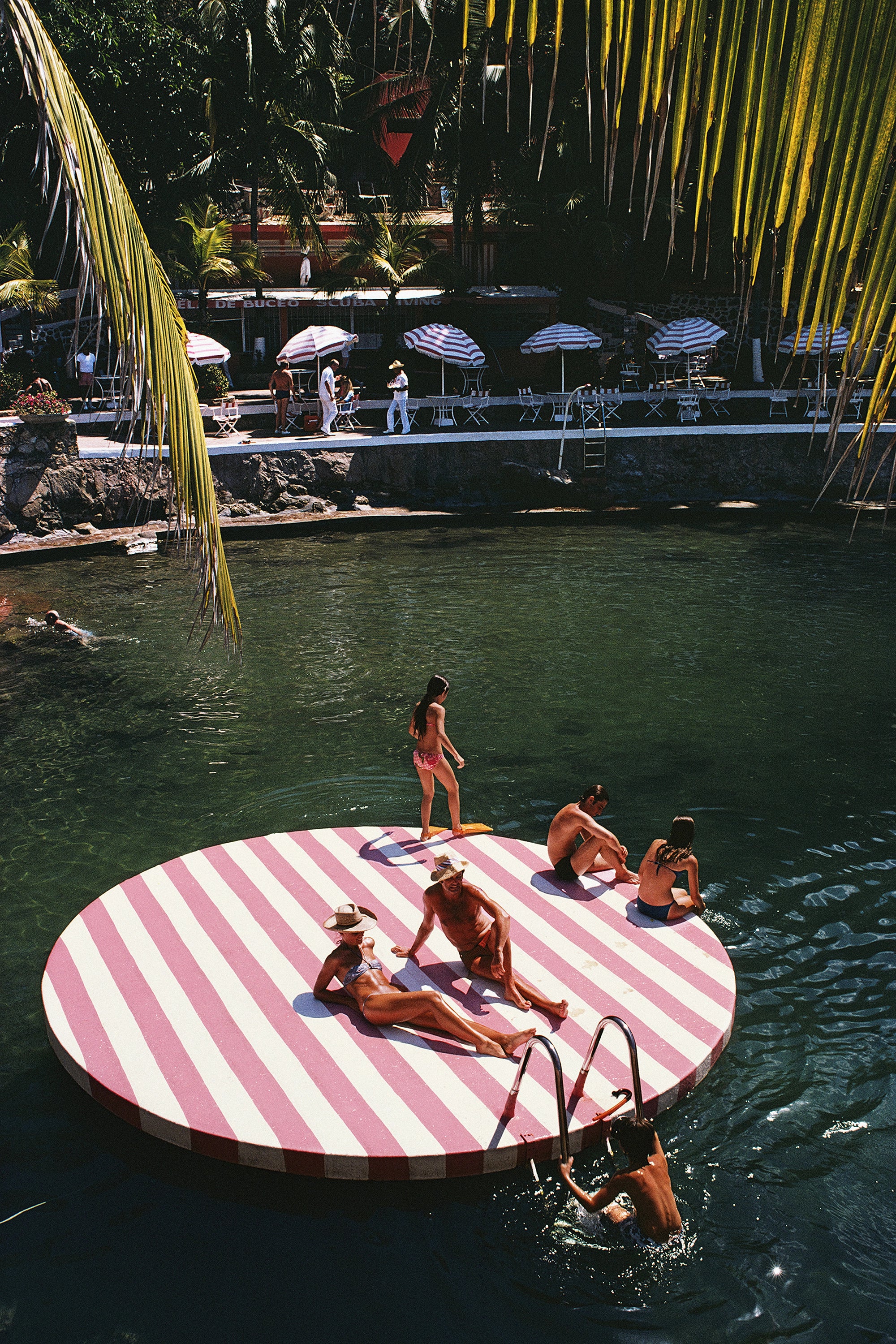 A large circular floating platform in a pool, featuring bold pink and white diagonal stripes. Several people are relaxing and standing on the platform, with palm leaves hanging over the scene and a resort-style background with umbrellas and lounge chairs.