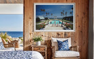 A framed art print depicting a tropical resort pool scene with palm trees and a blue sky, displayed on a rustic wooden wall above a wicker armchair in a styled bedroom setting. The Picturalist Slim Aarons Collection. 