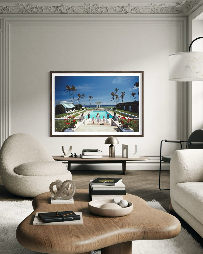 A framed art print depicting a luxurious tropical poolside scene with palm trees and a blue sky, displayed on a light gray wall in a modern, elegant living room setting. The Picturalist Slim Aarons Collection. 