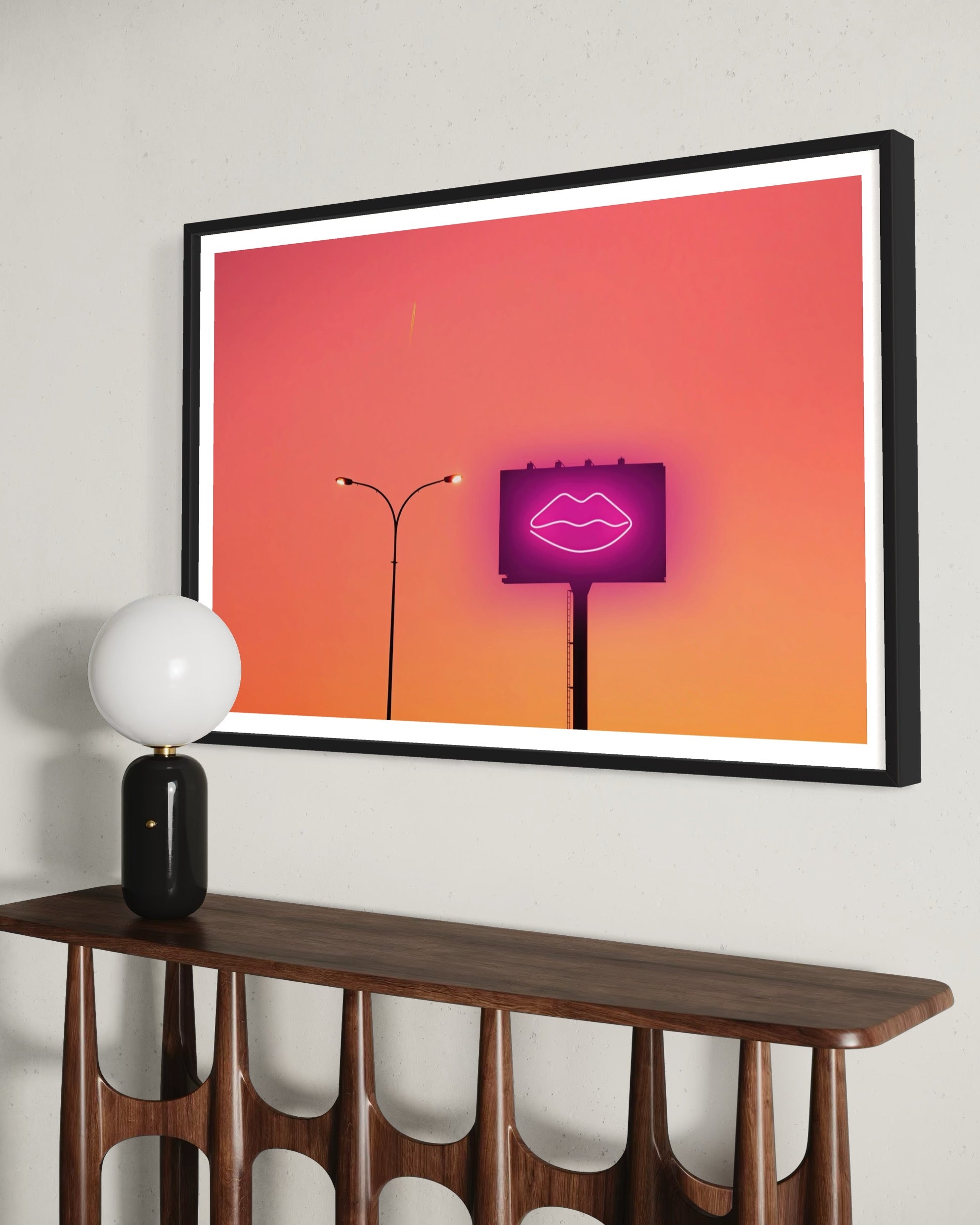 Framed artwork with a pink neon sign of lips on an orange background, displayed above a wooden console table.