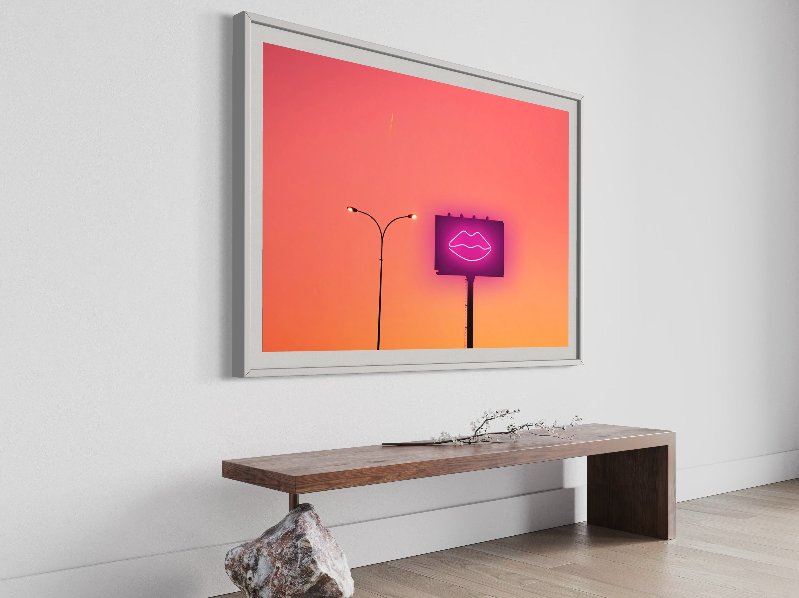 Framed artwork with a pink neon sign on a white wall above a wooden bench.
