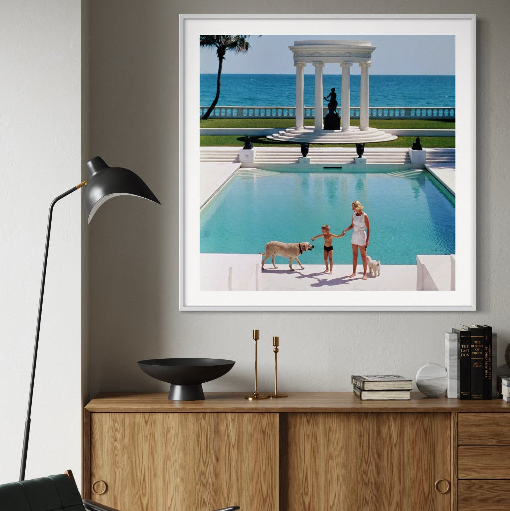Framed photograph of a pool scene with people and a dog, displayed on a wall above a wooden cabinet.