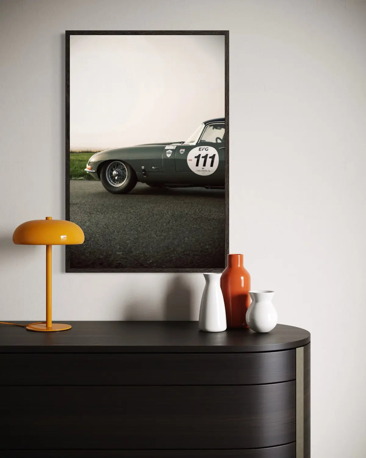A framed vertical art print of a vintage dark green race car on an asphalt road, styled on a light gray wall above a dark wood console table with a yellow lamp and decorative vases.
