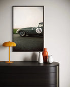 A framed vertical art print of a vintage dark green race car on an asphalt road, styled on a light gray wall above a dark wood console table with a yellow lamp and decorative vases.