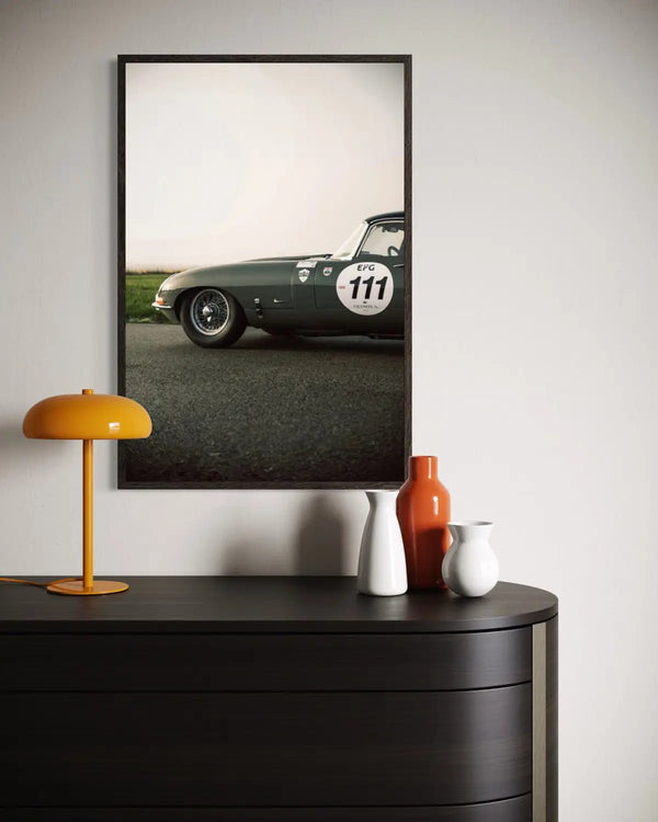 A framed vertical art print of a vintage dark green race car on an asphalt road, styled on a light gray wall above a dark wood console table with a yellow lamp and decorative vases.