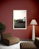 A framed vertical art print of a vintage green race car is displayed on a dark red wall in a modern living room setting with a brown leather lounge chair, a white floor lamp, and a potted plant.