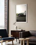 A framed vertical art print of a classic green race car with the number 111, displayed on a beige wall in a modern living room setting with a leather sofa, wooden sideboard, and blue armchair.