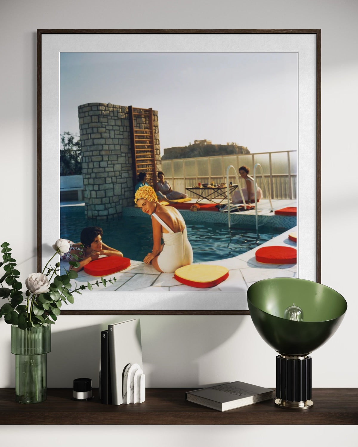 A framed art print depicting a vintage scene of people relaxing by a pool with red and yellow cushions, displayed on a shelf above a green bowl lamp and other decorative items.
