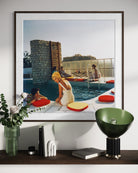 A framed art print depicting a vintage scene of people relaxing by a pool with red and yellow cushions, displayed on a shelf above a green bowl lamp and other decorative items.