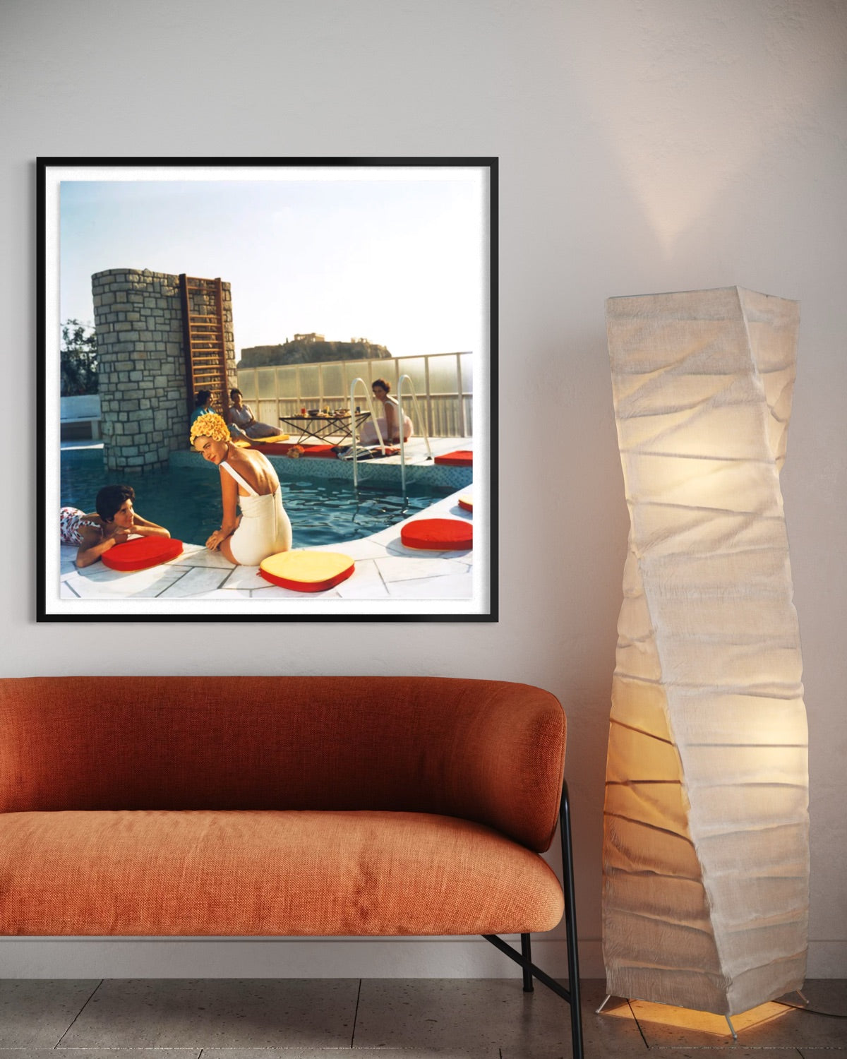A framed art print depicting a retro scene of women by a swimming pool with red float rings, displayed on a wall above an orange-brown velvet sofa next to a tall floor lamp.