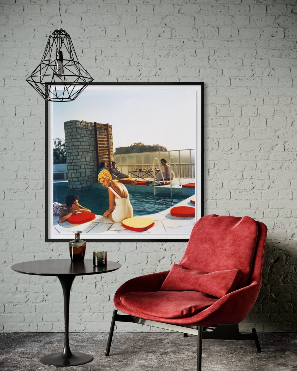 A framed art print depicting a retro scene of people relaxing by a swimming pool with red and yellow lounge chairs, styled on a white brick wall above a red armchair and a small black side table.