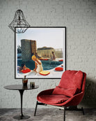 A framed art print depicting a retro scene of people relaxing by a swimming pool with red and yellow lounge chairs, styled on a white brick wall above a red armchair and a small black side table.