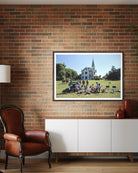 Framed picture of a group of people on a grassy field with a building in the background, displayed on a brick wall above a white cabinet.