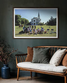 Framed photograph of a group of people on a grassy field with a large house in the background, displayed above a wooden bench with cushions.
