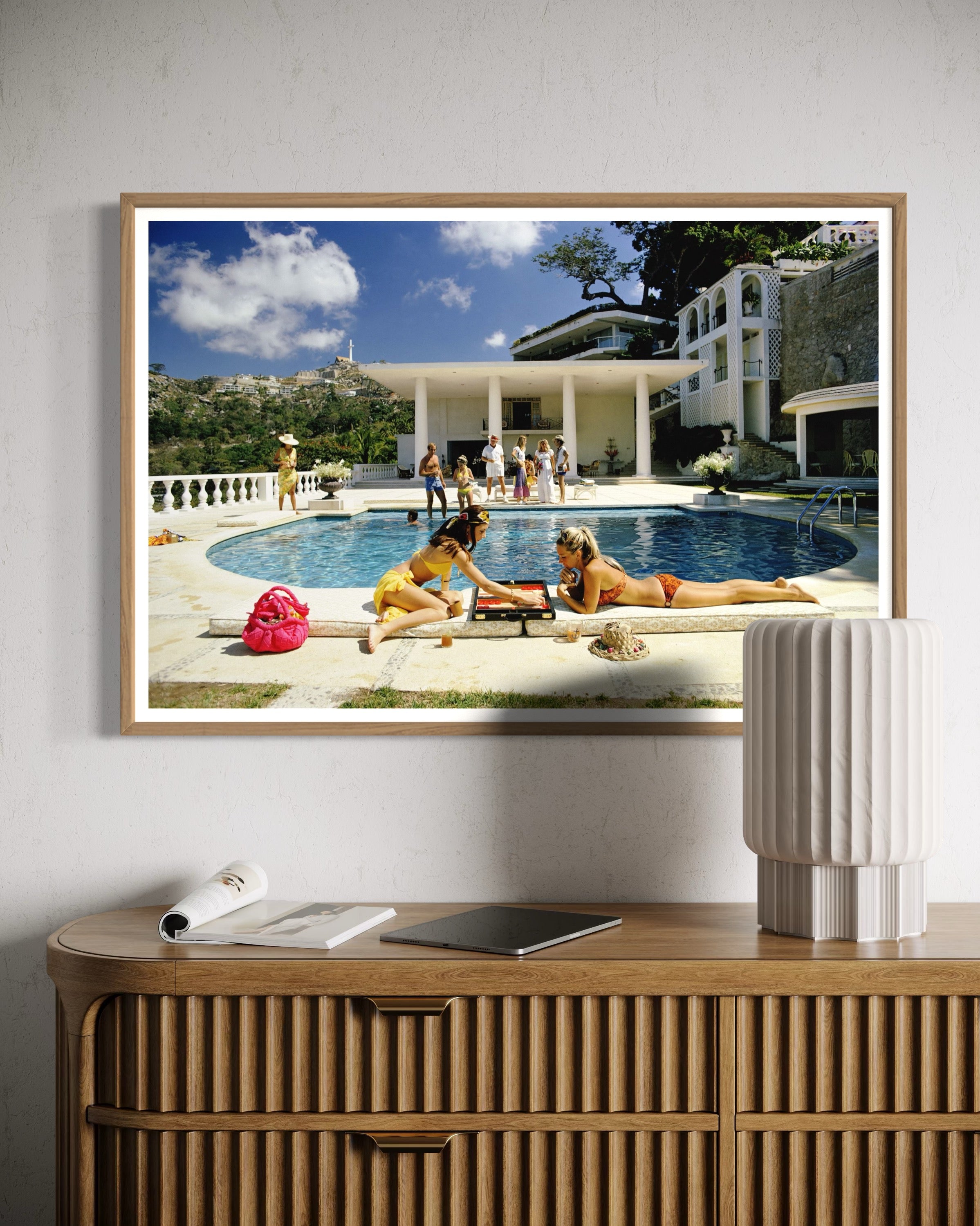Framed artwork of people by a pool on a wall above a wooden console table.
