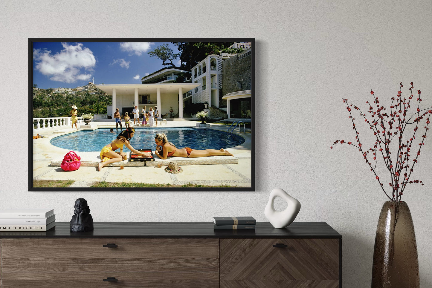 Mounted on a wall displaying an image of people by a pool, with decorative items on a wooden console table below.