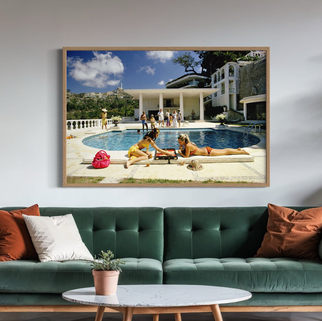 Framed artwork of people by a pool on a wall above a green sofa with decorative pillows.