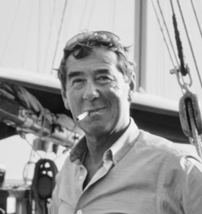 Black and white photo of a man with a pipe, wearing goggles on his head, standing in front of a boat.
