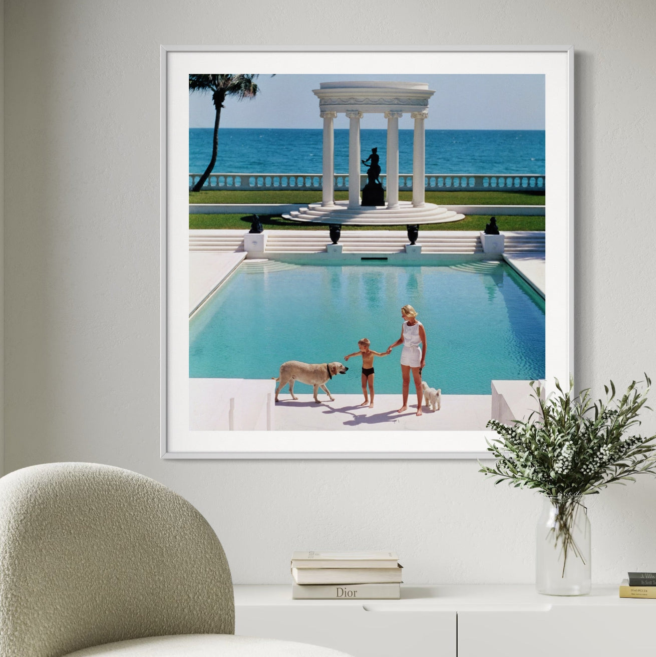 Framed photograph of a pool scene with people and dogs, displayed on a wall above a white cabinet.