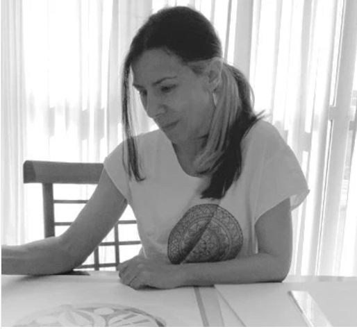 Woman sitting at a table with a decorative pillow on her lap, in a room with white curtains.