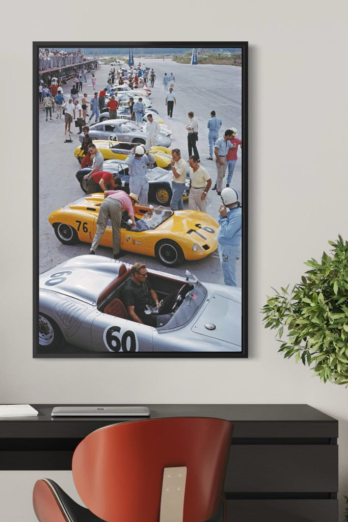 Framed artwork of a race car scene on a wall above a desk with a plant and lamp.