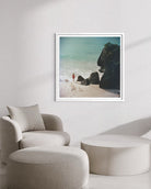A framed square art print depicting a beach scene with turquoise water, dark rocks, and pale sand, displayed on a light wall above a beige armchair and round ottoman.