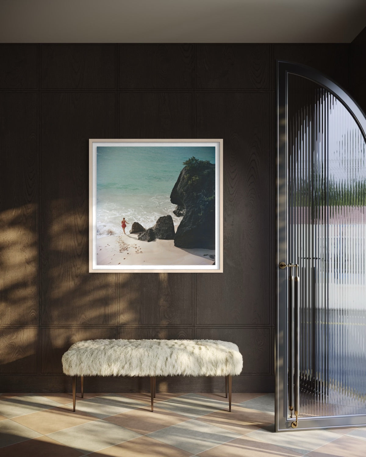 A framed square art print depicting a beach scene with turquoise water, dark rocks, and a small figure, displayed on a dark wood-paneled wall above a white faux-fur bench in a modern interior.