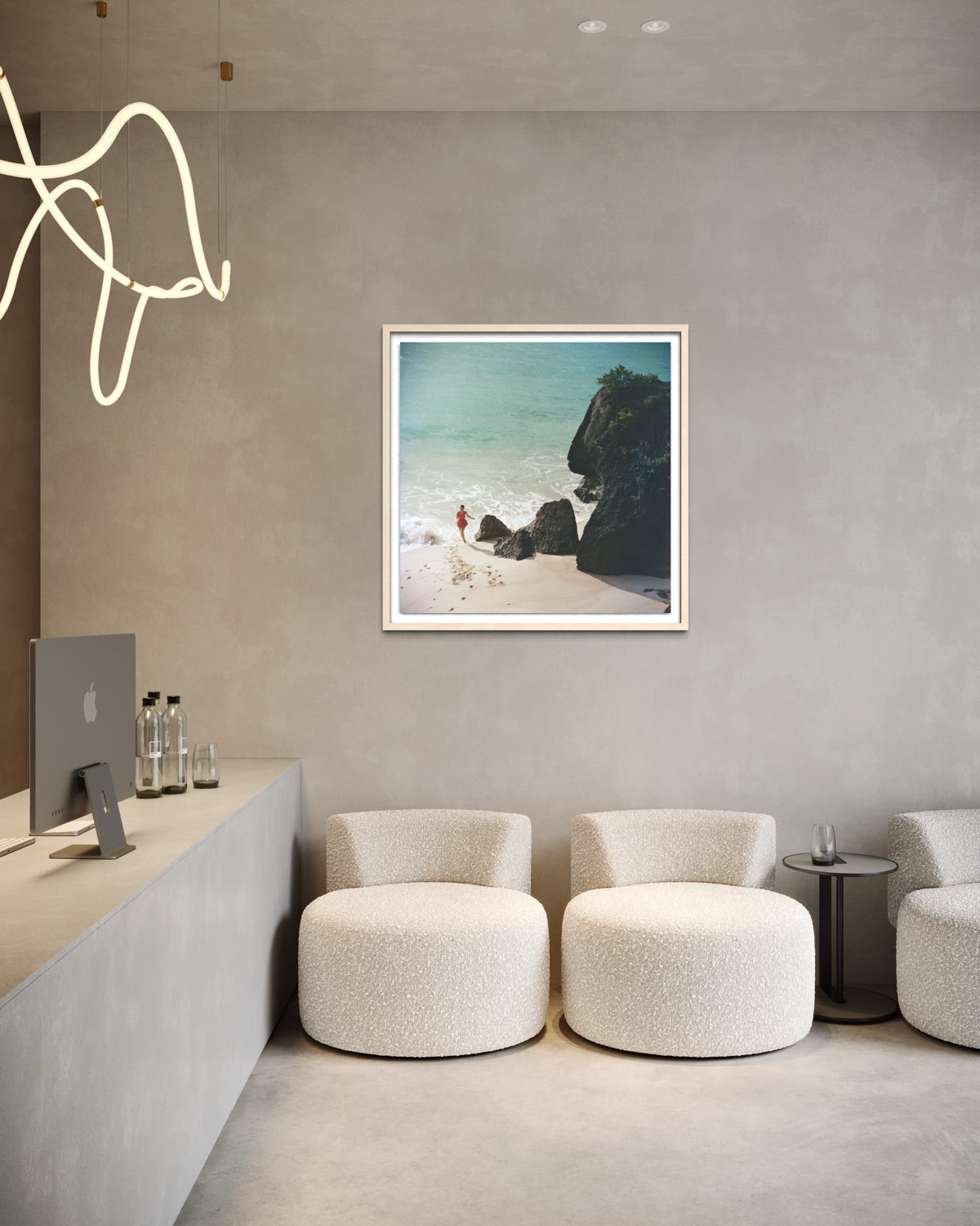 A framed square art print depicting a beach scene with turquoise water, white sand, and dark rocks, displayed on a textured gray wall in a modern minimalist interior with cream boucle chairs and a concrete counter.