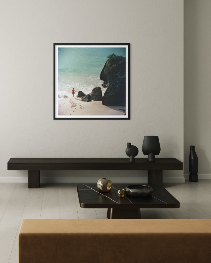 A framed square art print depicting a beach scene with turquoise water, white sand, dark rocks, and a small figure, displayed on a light gray wall above a dark wooden console table in a modern living room setting.