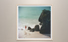 A framed square art print depicting a beach scene with turquoise water, white waves, dark rocks, and a small figure in red near the shoreline, mounted on a light beige wall.