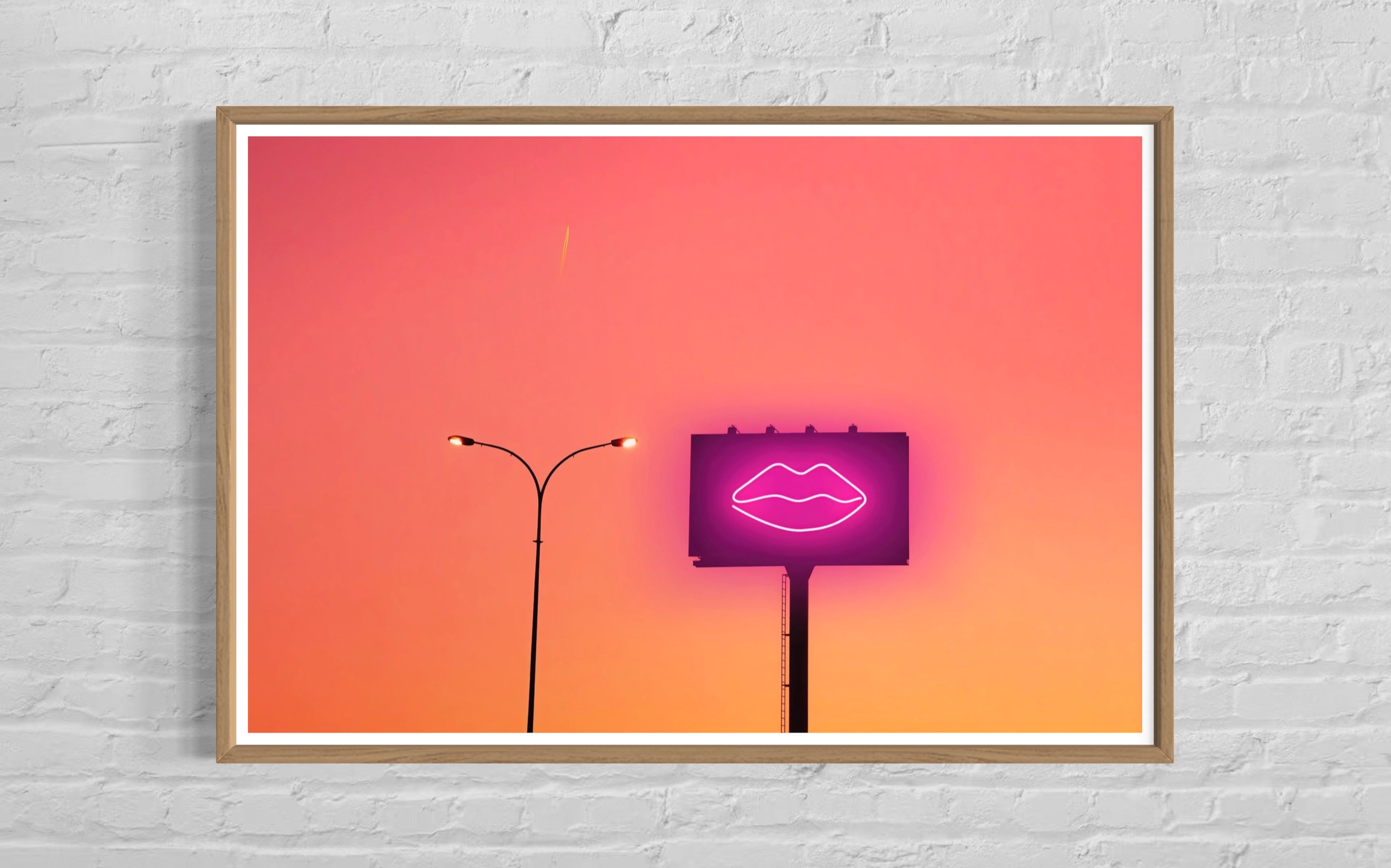 Framed artwork of a neon sign with lips against an orange and pink gradient sky.