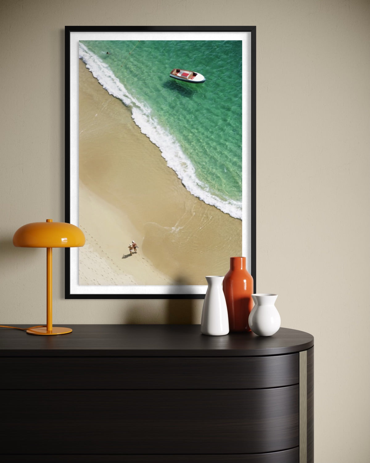 A framed aerial beach print hangs on a beige wall above a dark wood console table. The artwork shows turquoise water, white surf, and pale sand, with a small boat and tiny people visible.