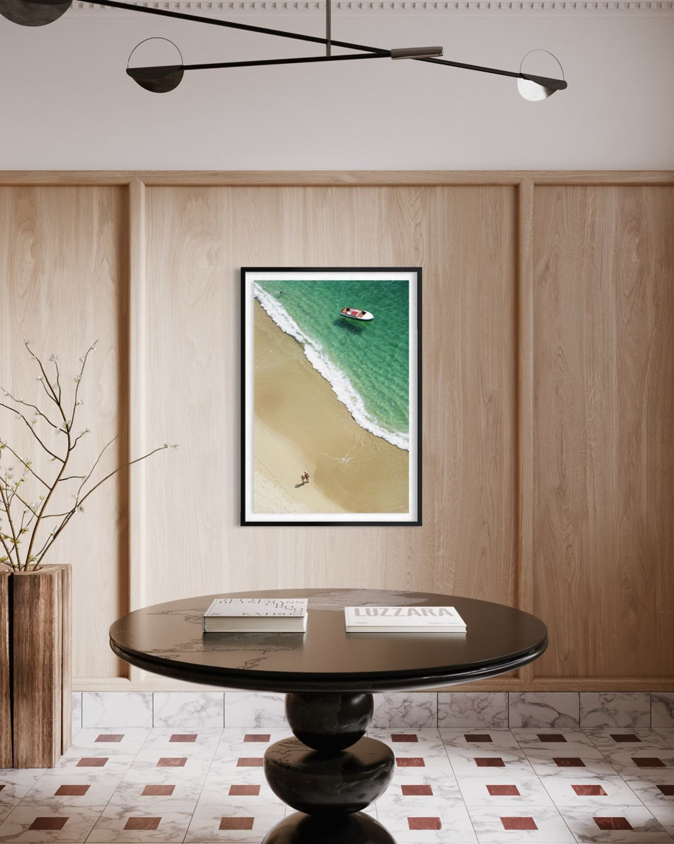 A framed vertical art print depicting an aerial view of a beach and turquoise ocean, displayed on a light wood paneled wall above a round black table in a modern interior setting.