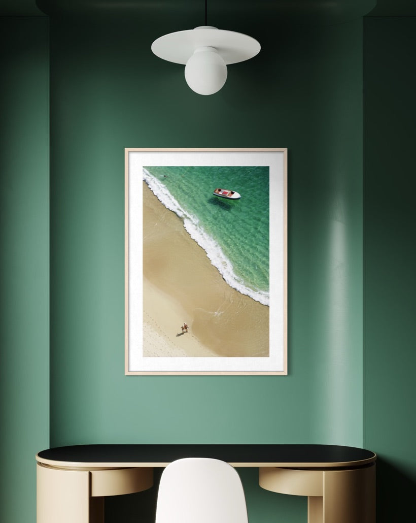 A framed vertical art print of an aerial beach scene with turquoise water and sand, displayed on a dark green wall above a desk in a modern interior setting.