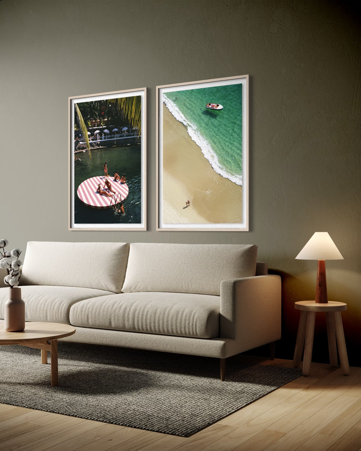 A modern living room scene with a light beige sofa, a small wooden side table with a lit lamp, and two framed beach-themed wall art prints hanging above the sofa. The prints show a red-and-white striped floatation pad and a turquoise ocean shoreline.