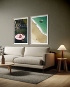 A modern living room scene with a light beige sofa, a small wooden side table with a lit lamp, and two framed beach-themed wall art prints hanging above the sofa. The prints show a red-and-white striped floatation pad and a turquoise ocean shoreline.