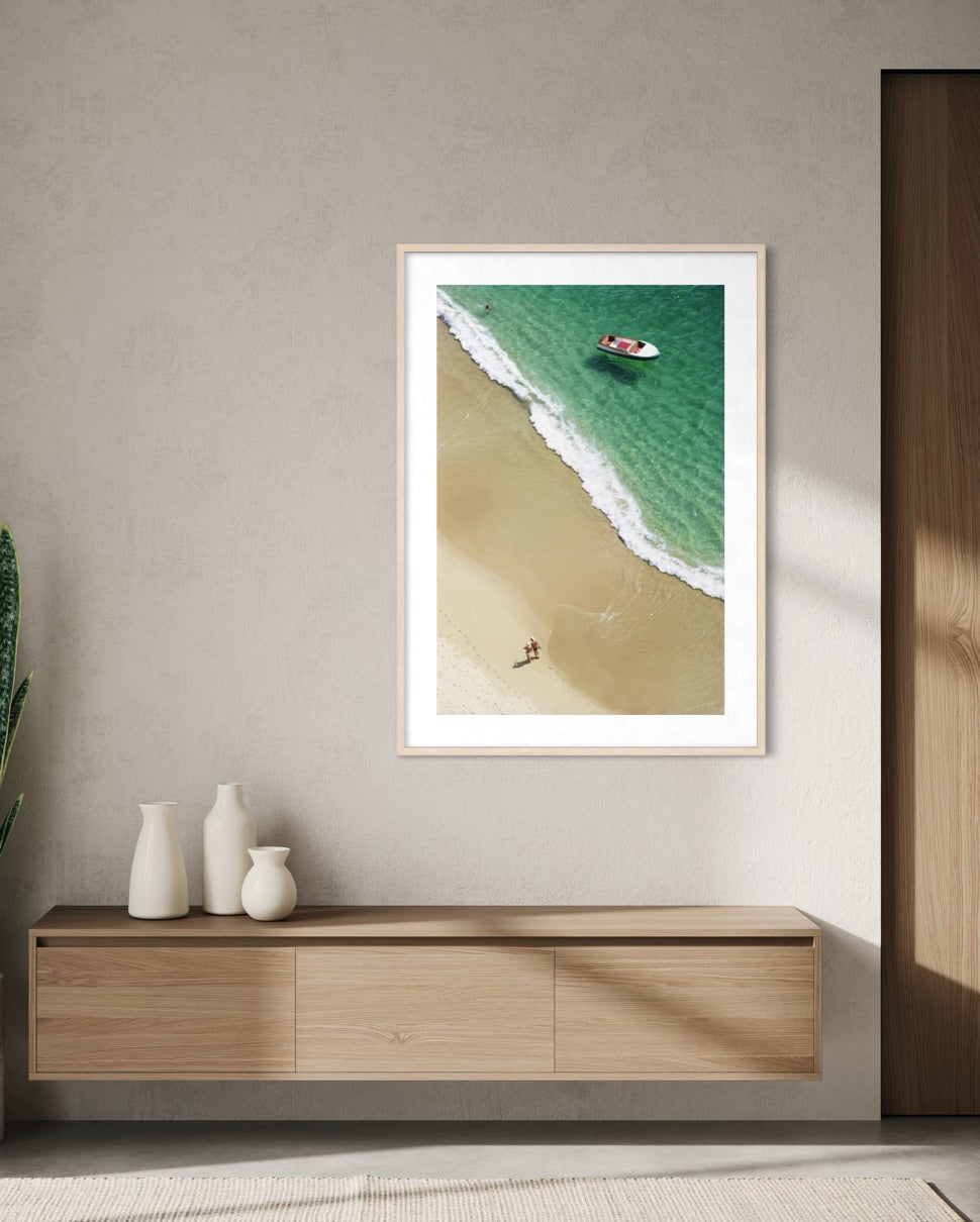 A framed vertical art print of an aerial beach scene with turquoise water, white surf, and pale sand, displayed on a light gray wall above a wooden console with white vases.