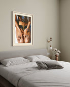 A framed art print featuring a close-up photograph of a person wearing a black bikini top with a large blue gem and a gold cross necklace, displayed on a beige wall above a bed in a modern bedroom setting.