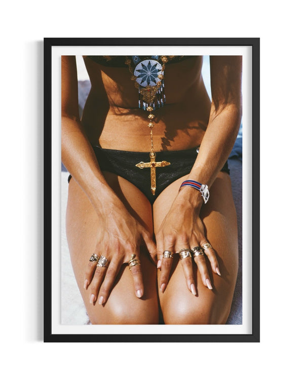 A framed art print featuring a close-up photograph of a woman wearing black bikini bottoms and multiple gold rings, with a black frame and white mat on a white background.